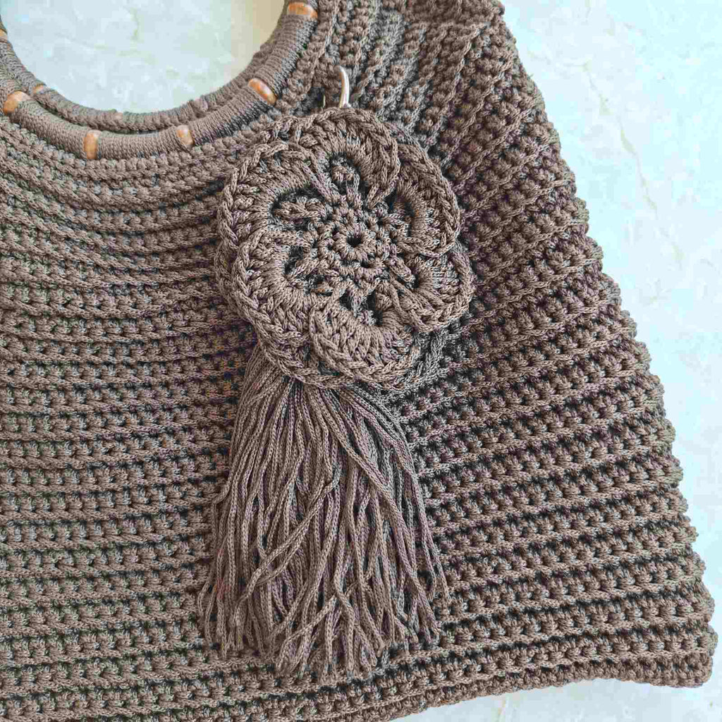 A close-up of a brown crocheted shoulder bag featuring a large decorative crocheted flower applique with a long tassel on the front, set against a light textured background.Detail of hanging flower on handmade crochet bag – unique artisan accessory Georgia Create