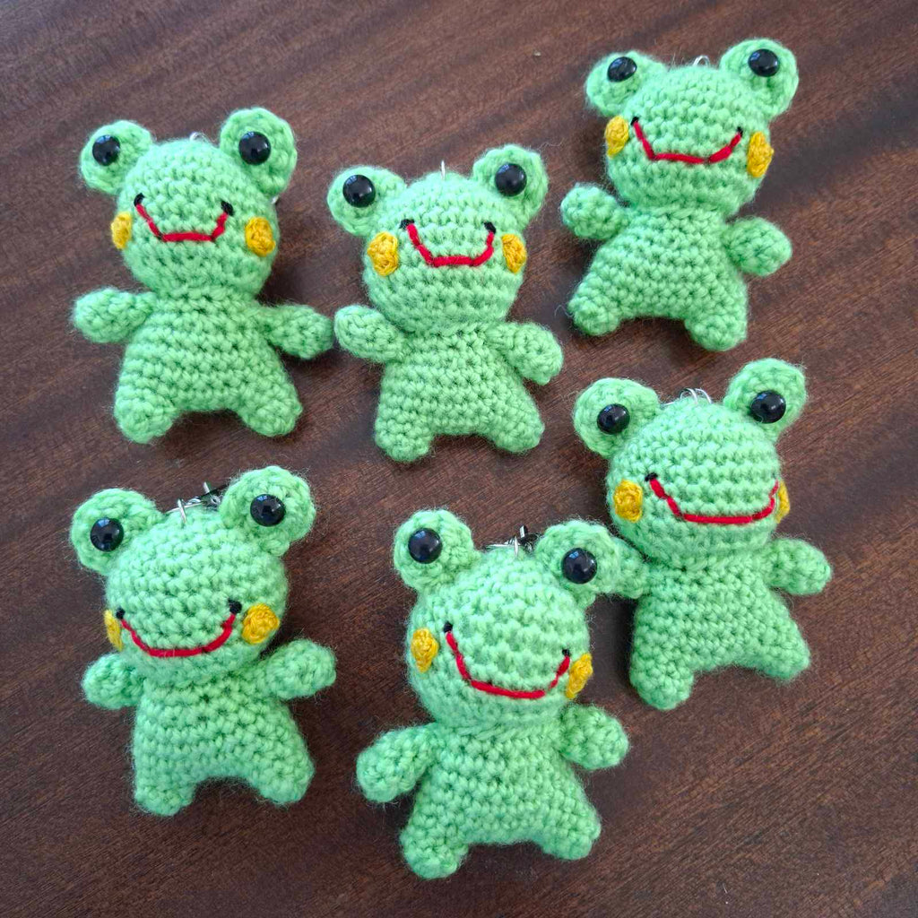 Seven handmade crochet frog keychains laid flat on a wooden surface