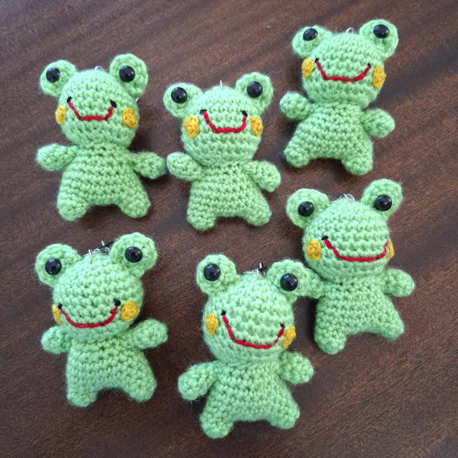 Seven handmade crochet frog keychains laid flat on a wooden surface