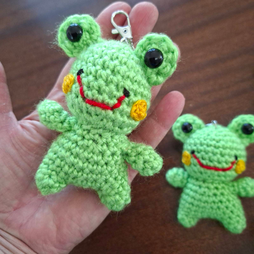 Handmade crochet frog keychain held in hand showing size, with a second frog in the background