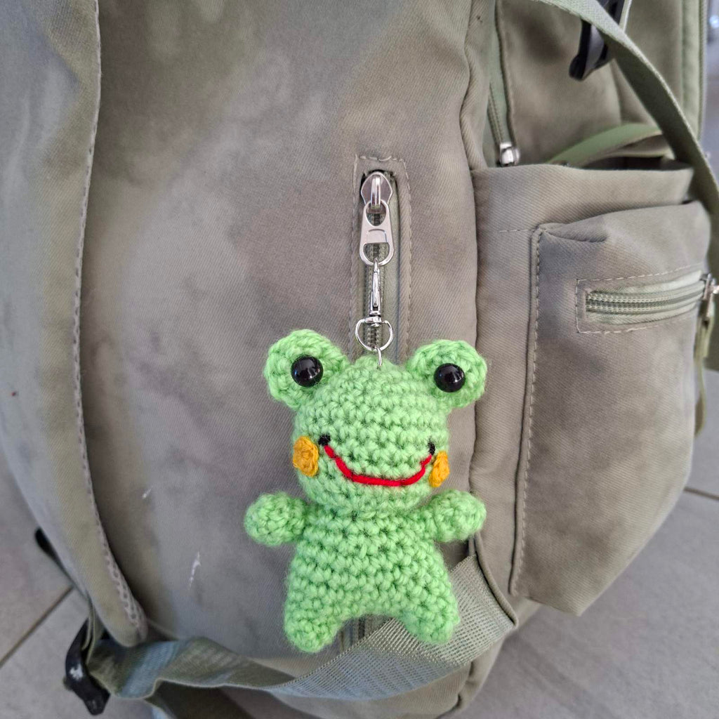 Crochet frog keychain clipped onto a grey backpack zipper