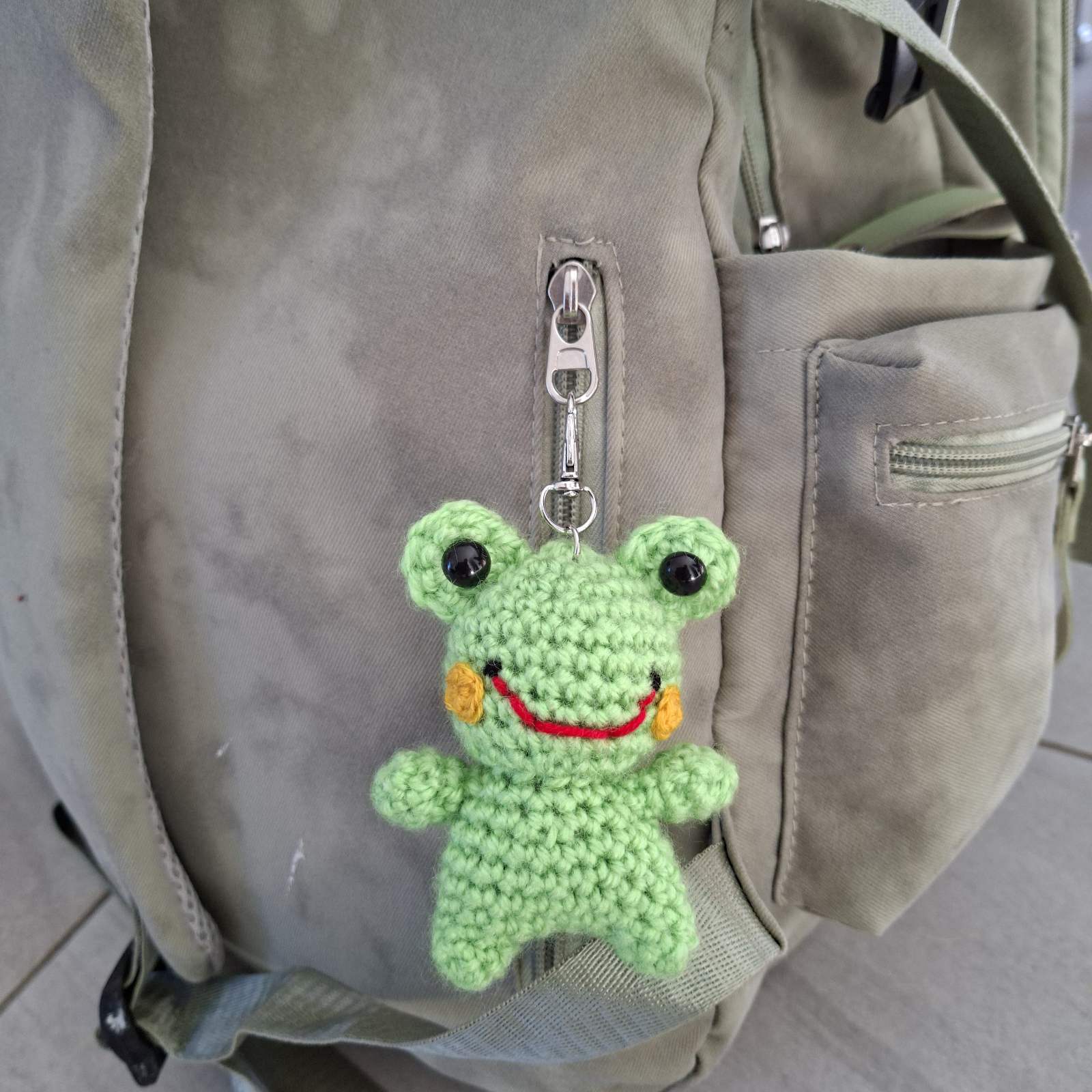 Crochet frog keychain clipped onto a grey backpack zipper
