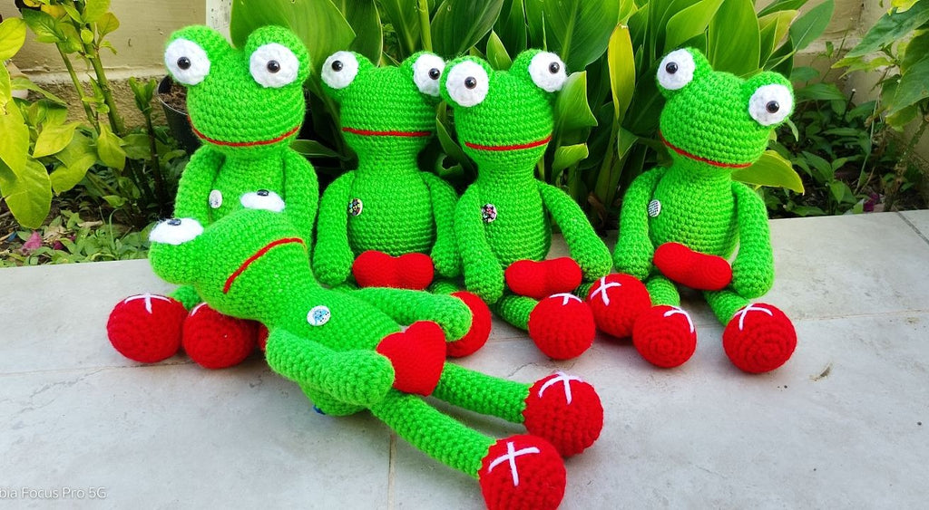 Five green crocheted frog toys with large white eyes and red hearts are arranged on a light stone surface with green plants in the background.