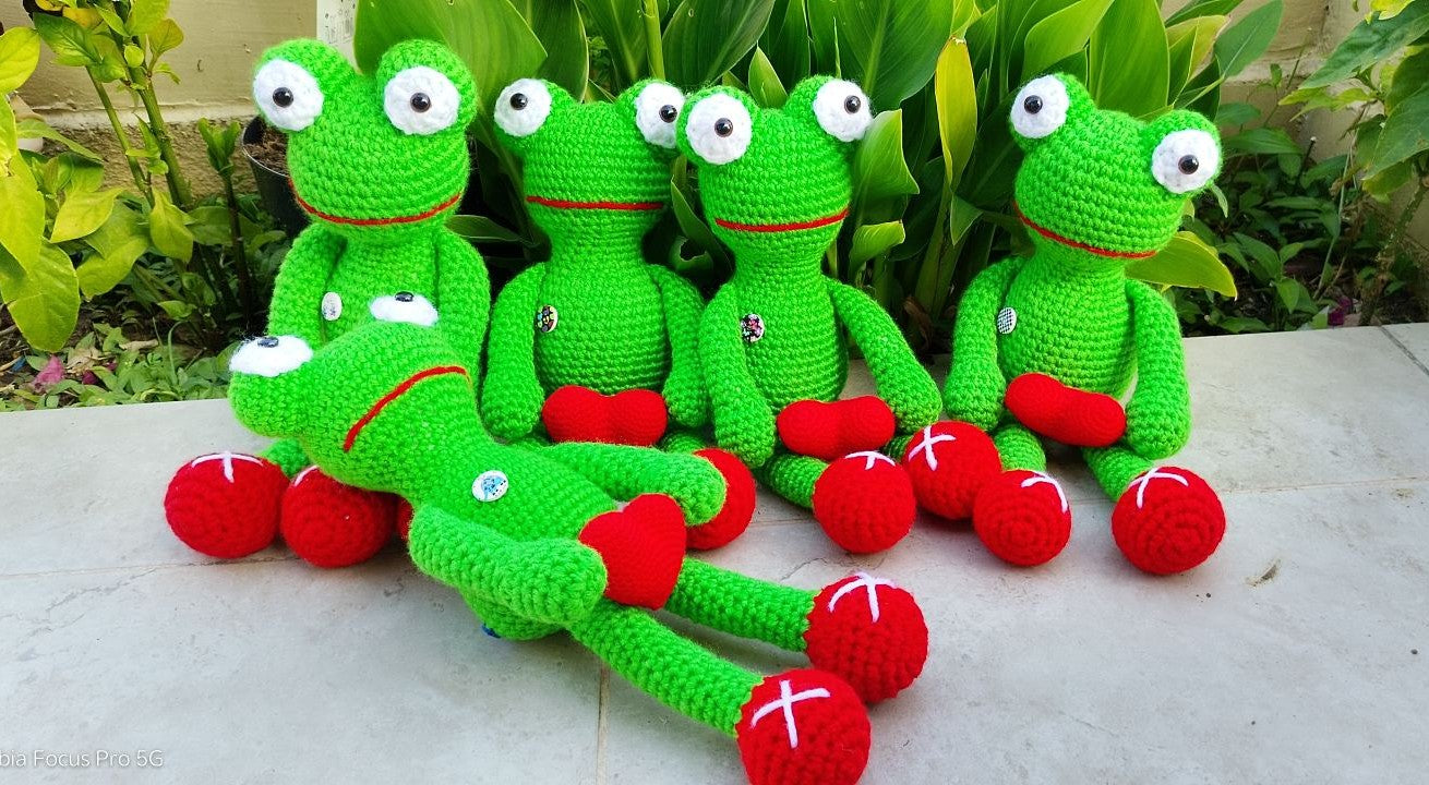 Five green crocheted frog toys with large white eyes and red hearts are arranged on a light stone surface with green plants in the background.