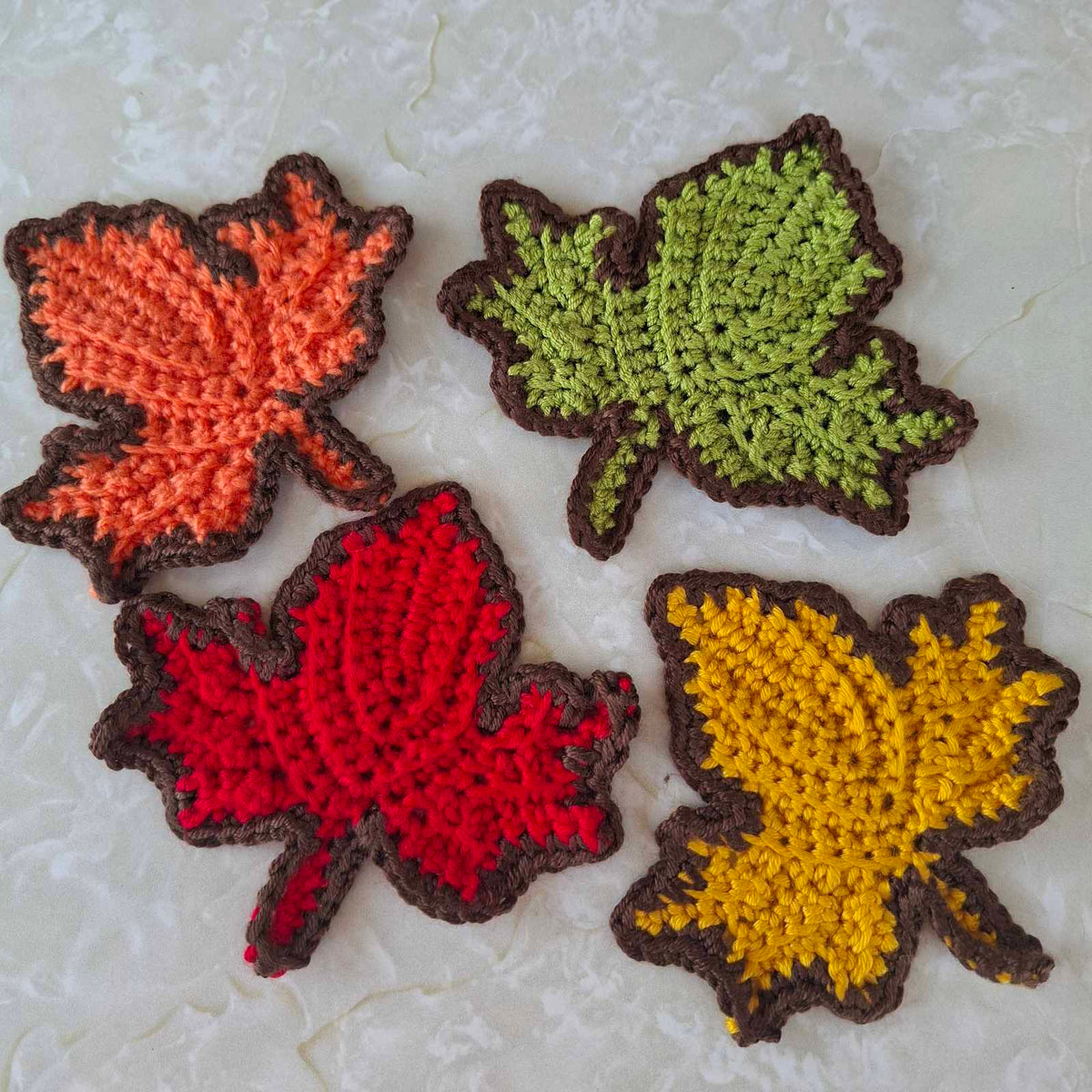 Set of four crochet autumn leaf coasters in orange, green, red and mustard yellow with brown edging